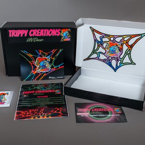Pu&ograve; includere: Una scatola nera con la scritta "Trippy Creations UV Decor" sul davanti. La scatola &egrave; aperta e contiene un adesivo colorato con un personaggio dei cartoni animati. L'adesivo &egrave; su uno sfondo bianco. Ci sono anche altri due adesivi con lo stesso personaggio.