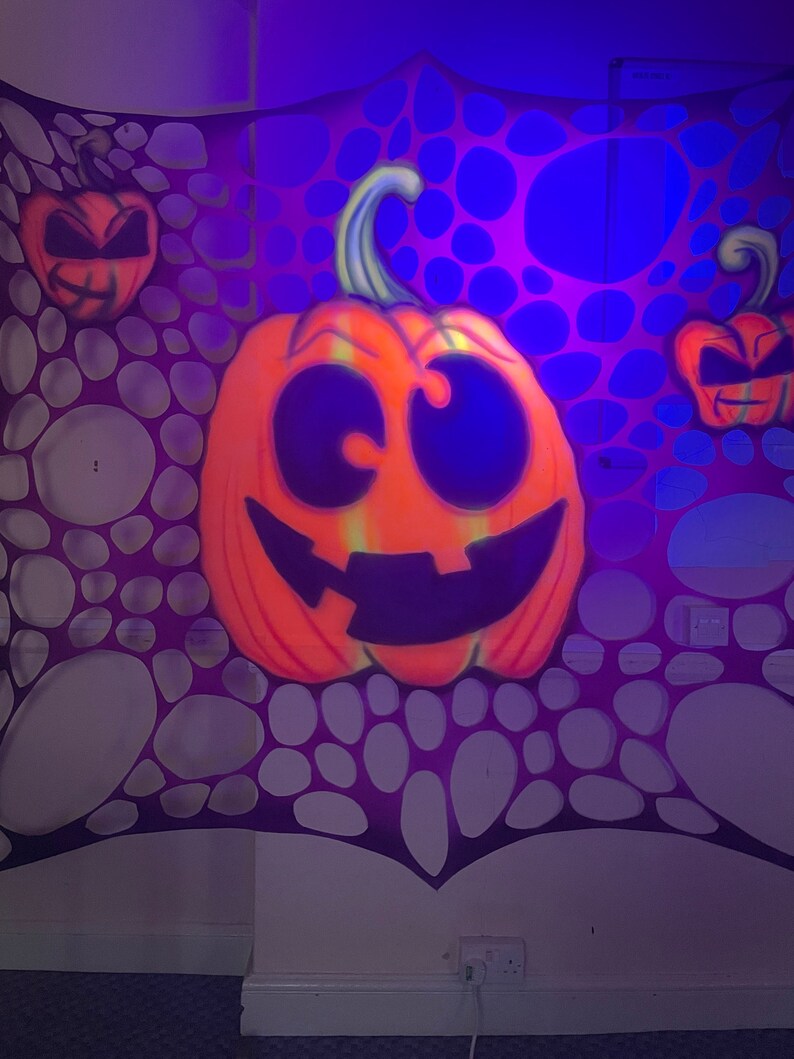 Pu&ograve; includere: Una decorazione da parete a tema Halloween con una grande zucca arancione con un viso bianco e nero e uno sfondo di ragnatela viola e bianco.