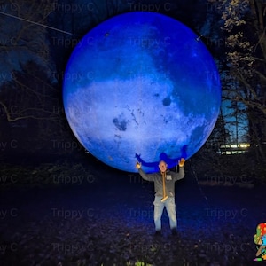 Op de afbeelding: Een grote, lichtgevende, bolvormige installatie die lijkt op een blauwe maan, hangend in een donkere buitenomgeving. Het oppervlak van de bol vertoont getextureerde, kraterachtige patronen in verschillende tinten diepblauw en lichtblauw.