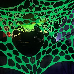 Könnte beinhalten: Eine neongrüne, netzartige Struktur mit großen, unregelmäßigen Löchern. Die Struktur ist beleuchtet und erzeugt einen Leuchteffekt. Im Hintergrund ist eine Person zu sehen. Die Szene ist dunkel, was auf eine Party oder Veranstaltung hindeutet.
