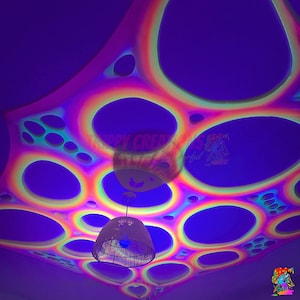 May include: A ceiling canopy featuring a vibrant, multi-colored rainbow gradient pattern with large, circular cutouts of varying sizes. The fabric displays a glowing effect under ultraviolet light, and a woven pendant light fixture hangs centrally.