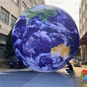 Op de afbeelding: Een grote opblaasbare wereldbol die de aarde afbeeldt, met groene en gele continenten tegen een blauwe oceaan. De wereldbol staat op een grijze ondergrond, met een persoon die in de buurt knielt. De foto is buiten genomen, met gebouwen op de achtergrond.