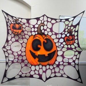 Pu&ograve; includere: Una ragnatela viola e nera con tre zucche arancioni con occhi e bocche nere. Le zucche sono su un muro bianco.