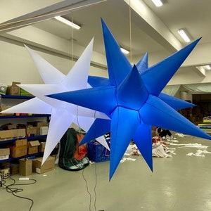 Op de afbeelding: Twee grote, opblaasbare sterdecoraties, één wit en één blauw, hangend aan het plafond. De sterren hebben meerdere punten en zijn waarschijnlijk bedoeld voor feestelijke gelegenheden. De achtergrond is goed verlicht, met zichtbare snoeren.