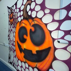 Pu&ograve; includere: Una zucca arancione intagliata con un viso sorridente nero &egrave; visibile attraverso uno schema di ragnatela viola e bianca.