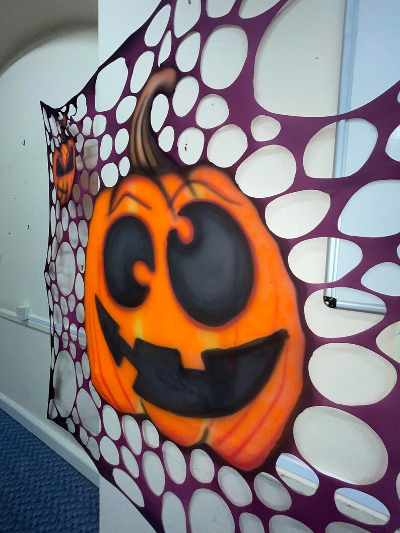 Pu&ograve; includere: Una zucca arancione intagliata con un viso sorridente nero &egrave; visibile attraverso uno schema di ragnatela viola e bianca.