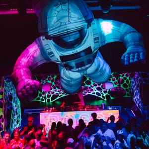 May include: An inflatable astronaut figure hovers above a crowd at a nightclub. The astronaut is primarily light blue with pink and purple lighting. The stage features a DJ setup and the word "POSS" in colorful letters. The walls have neon green and black patterns.