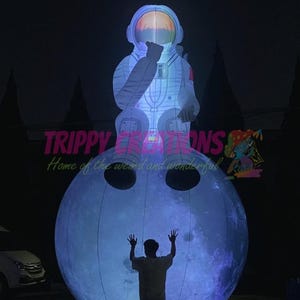 Op de afbeelding: Een verlichte opblaasbare astronautfiguur zittend op een blauwe maan, met een persoon eronder met de armen omhoog. De astronaut draagt een wit ruimtepak. De tekst "TRIPPY CREATIONS Home of the weird and wonderful" is zichtbaar.