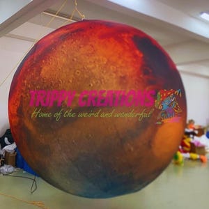 Op de afbeelding: Grote opblaasbare bol met een gedetailleerd Mars-oppervlakontwerp, met tinten rood, oranje en bruin. De bol heeft de tekst "TRIPPY CREATIONS" en "Home of the weird and wonderful".