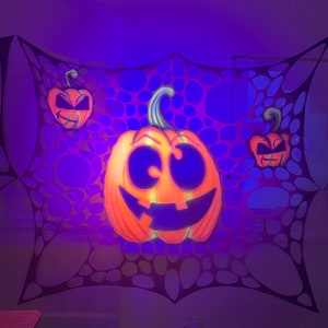Pu&ograve; includere: Tre zucche arancioni a forma di jack-o'-lantern con occhi e bocche nere sono appese a una ragnatela nera su uno sfondo viola.