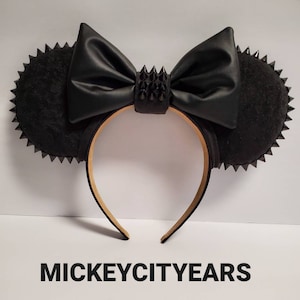 May include: Black mouse ear headband with a large bow and spiked accents. The ears are round and have a spiked trim. The bow and spikes are black. The headband is a light brown color. The text "MICKEYCITYEARS" is at the bottom.