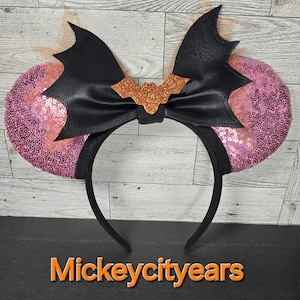 May include: Pink and black Minnie Mouse ears with a black bat bow and orange glitter accents. The ears are covered in pink sequins. The bow has a black bat silhouette in the center.