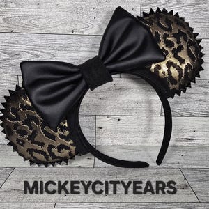 May include: Black and gold leopard print Minnie Mouse ears with a black satin bow. The ears have a spiky design around the edges.