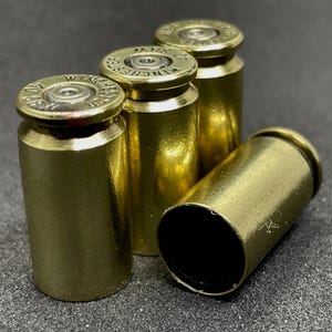 Peut inclure: Gros plan sur quatre douilles en laiton, dont trois sont dressées et une sur le côté. Les douilles ont un aspect doré brillant avec des marquages visibles sur le dessus. Le fond est une surface sombre et texturée.