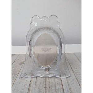 May include: Clear, decorative photo frame with a floral design. The frame has an oval opening for a photo and is made of clear plastic. The frame is on a light-colored wooden surface.