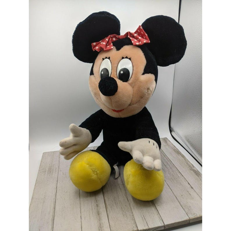 May include: A plush black and brown Minnie Mouse toy with a red bow with white polka dots. The toy is sitting with its legs crossed and has yellow feet.