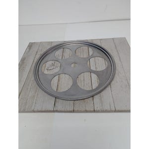 Op de afbeelding: Een cirkelvormige, zilverkleurige metalen eierhouder met zeven ronde uitsparingen. De eierhouder ligt op een verweerd, lichtgrijs houten oppervlak. Het metaal heeft een matte afwerking.