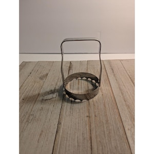 May include: A circular metal biscuit cutter featuring a scalloped edge and a tall, arched handle. The tool is constructed from silver-toned metal with a smooth finish, designed for cutting dough into uniform rounds with decorative edges.