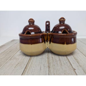 May include: A two-section ceramic condiment server with matching lids and a handle. The server is two-toned, with a brown top and a tan bottom. The lids have a small spoon rest. The server is on a wooden surface.