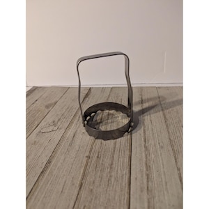 May include: A silver-colored metal biscuit cutter featuring a circular base with a scalloped edge and a tall, arched handle attached to the sides. The tool is constructed from a single piece of bent metal, providing a sturdy grip for cutting dough.