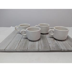 May include: Four small, white ceramic coffee cups with rounded handles are arranged on a weathered, gray wooden tray. The cups are evenly spaced, showcasing their simple, classic design. The background is a plain white surface.