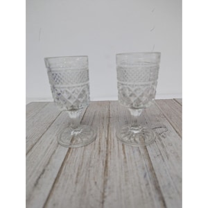 May include: Two clear, vintage-style stemmed glasses with diamond-patterned detailing. The glasses sit on a light-colored, weathered wooden surface. The glasses have a rounded base and a short stem.