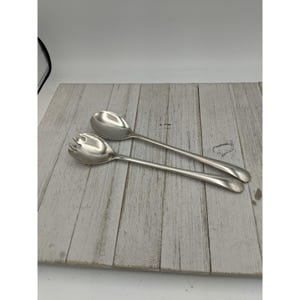 May include: A set of two silver salad serving utensils, a spoon and a fork, on a white wooden surface.