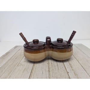 May include: A two-section ceramic condiment server with a handle, lids, and wooden spoons. The server is brown and tan, with a handle in the middle. The lids and spoons are dark brown. The server is on a light wood surface.