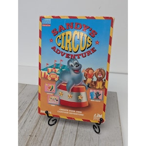 May include: A vintage "Sandy's Circus Adventure" interactive CD-ROM game box. The box features a blue background with a cartoon seal, circus tent, and the text "Sandy's Circus Adventure". The box is displayed on a black metal stand.