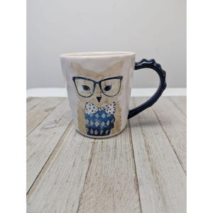 Puede incluir: Una taza de cerámica color crema con un diseño de búho caprichoso. El búho lleva gafas y una pajarita con un estampado azul y blanco. El asa es azul oscuro. La taza tiene una superficie ligeramente texturizada.