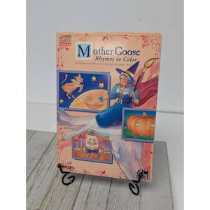 May include: A vintage "Mother Goose Rhymes to Color" book. The cover features illustrations of nursery rhyme characters, including a woman with a paintbrush, a cow jumping over the moon, and Humpty Dumpty. The book is displayed on a black metal stand.