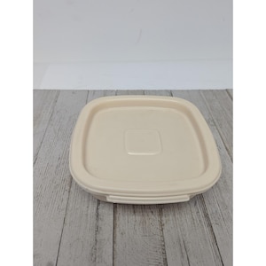 Puede incluir: Una tapa cuadrada de plástico blanquecino con un centro empotrado y un borde ligeramente elevado. La tapa está diseñada para encajar en un recipiente y se muestra sobre una superficie de madera clara.