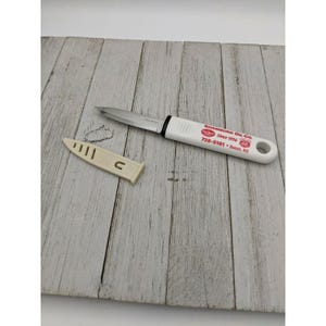 May include: A stainless steel paring knife with a white handle and a beige sheath. The handle has red text that reads "Shamburg Oil Co. Since 1936" and the number "66". The knife is on a white wooden surface.