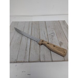 May include: A stainless steel knife with a wooden handle. The knife is resting on a weathered, light gray wooden surface. The handle is light brown with three rivets.