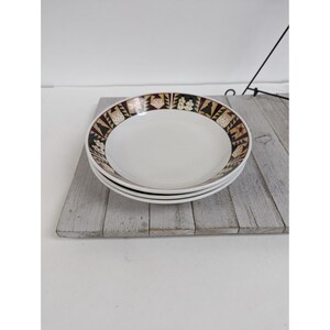 May include: A stack of three white ceramic bowls with a decorative border. The border features a repeating pattern of geometric shapes in brown, green, and gold against a black background. The bowls are stacked on a weathered wooden surface.