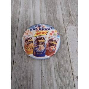 Puede incluir: Un botón redondo azul y blanco con una imagen de tres cajas de cereal Snack Ums. Las cajas son roja, amarilla y azul. El texto en el botón dice "ask me about NEW! Snack Ums".