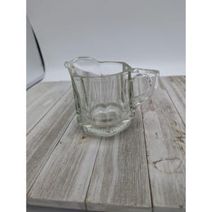 May include: A clear glass creamer with a handle. The creamer has a faceted design and is perfect for serving cream or milk.