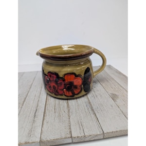 Puede incluir: Jarra de cerámica con asa, con diseño floral en rojo y negro sobre fondo beige. La jarra tiene un borde marrón y un pico vertedor. Probablemente sea un artículo vintage o hecho a mano.