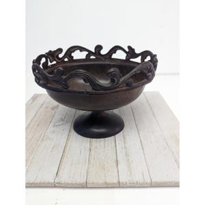 Puede incluir: Un cuenco decorativo marrón oscuro con base de pedestal. El borde del cuenco presenta un diseño ornamental y ondulado. El cuenco es de metal y se asienta sobre una superficie de madera clara.