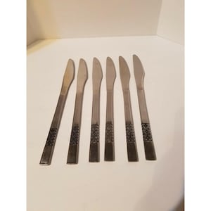 May include: Set of six stainless steel knives. Each knife has a long, slender blade and a handle with a decorative, embossed pattern. The knives are arranged on a white surface.