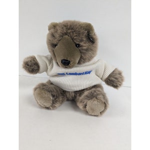 May include: A brown teddy bear wearing a white sweater with blue lettering that says "The Laundry".