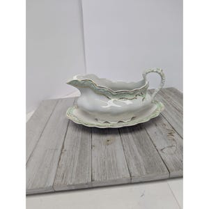 Puede incluir: Una salsera de porcelana blanca con plato integrado, que presenta un borde festoneado y un delicado patrón de filigrana en verde claro y dorado a lo largo de los bordes. La pieza incluye un asa curva y un pico vertedor texturizado.