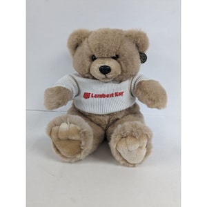 May include: A brown teddy bear wearing a white sweater with the red text "Lambert Kay" on the front.