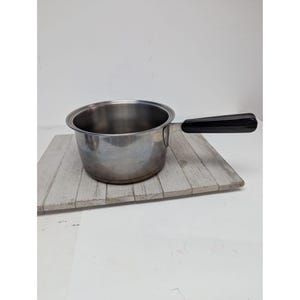May include: A stainless steel saucepan with a black handle. The pot is empty and sits on a light grey wooden surface. The handle is angled and appears to be made of a hard plastic material. The pot is round with a slightly flared rim.