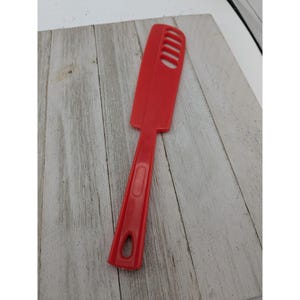 May include: A bright red spatula with a long handle and a rectangular head with several slots. The spatula is made of a solid, smooth material and is designed for kitchen use. The handle has a hole for hanging.