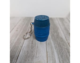 Puede incluir: Llavero de barril de plástico azul con un anillo de metal. El barril tiene un diseño en relieve de un barril con las palabras "Old Smugglers" en él.