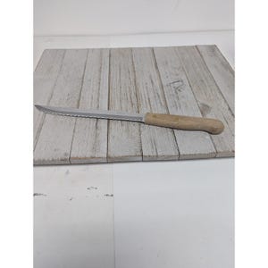 May include: A serrated bread knife with a wooden handle rests on a weathered, gray wooden cutting board. The stainless steel blade has a series of teeth along the cutting edge. The handle is light brown and appears to be made of wood.