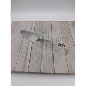 Puede incluir: Una espátula para pastel de acero inoxidable con una hoja ancha y plana y un mango largo. El mango presenta un diseño floral decorativo cerca del extremo. La espátula está sobre una superficie de madera clara.