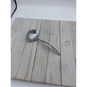 Puede incluir: Un cucharón de acero inoxidable con un cuenco redondo y un mango largo. El mango tiene un diseño texturizado a lo largo del borde superior. El cucharón está sobre una superficie de madera clara.