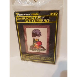 May include: A vintage crewel embroidery kit featuring a boy wearing a purple hat and holding a red helmet. The kit includes a pre-printed canvas, embroidery floss, and instructions. The kit is labeled "Vogart Crafts Moppets Crewel Creative Stitchery Kit" and "2460".